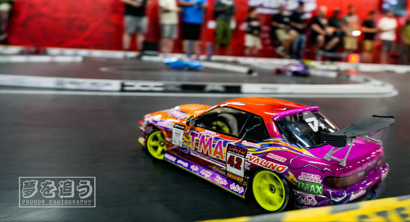 A drift racing car with a vibrant purple, orange, and pink livery and yellow wheels, captured in motion on a racetrack with blurred spectators in the background.